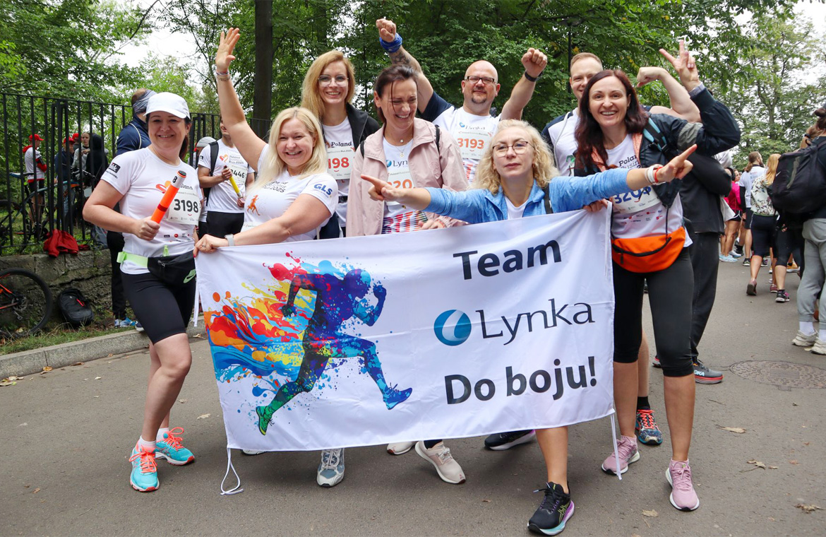 Uśmiechnięta drużyna Lynka w koszulkach startowych pozuje z banerem &bdquo;Team Lynka &ndash; Do boju!" po biegu.