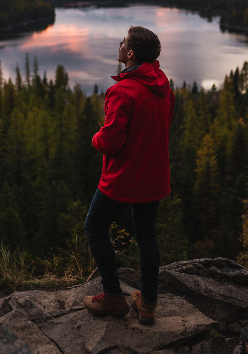 Man in a red softshell jacket looking out over a forest at sunset.